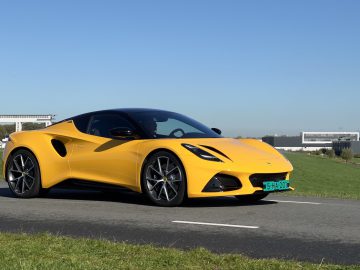 Een gele Lotus Emira met zwarte accenten staat geparkeerd op een weg met groen gras aan de zijkanten, onder een helderblauwe lucht.