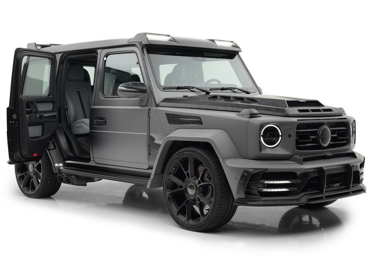 Een matzwarte luxe SUV van Mansory G-Klasse met de achterpassagiersdeur open, met een donker lederen interieur en op maat gemaakte zwarte velgen op een witte achtergrond.