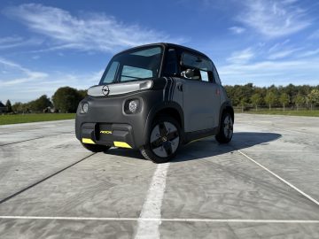 Een compacte, tweedeurs elektrische Opel Rocks-e-auto staat buiten op een betonnen ondergrond geparkeerd onder een helderblauwe lucht, met groene bomen op de achtergrond.