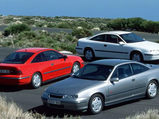 Drie vintage sportwagens, waaronder een Opel Calibra, een rode, een witte en een zilveren, staan geparkeerd op een asfaltweg met groen en heuvels op de achtergrond.