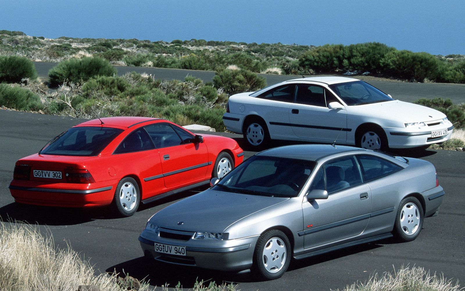 Drie vintage sportwagens, waaronder een Opel Calibra, een rode, een witte en een zilveren, staan geparkeerd op een asfaltweg met groen en heuvels op de achtergrond.