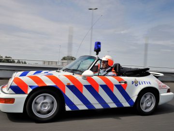 Een persoon die in een witte politie-Porsche met blauwe en rode strepen en een blauwe sirene rijdt, snelt over de snelweg. De bestuurder, gekleed in een witte helm, lijkt gefocust op het uitdelen van verkeersboetes.