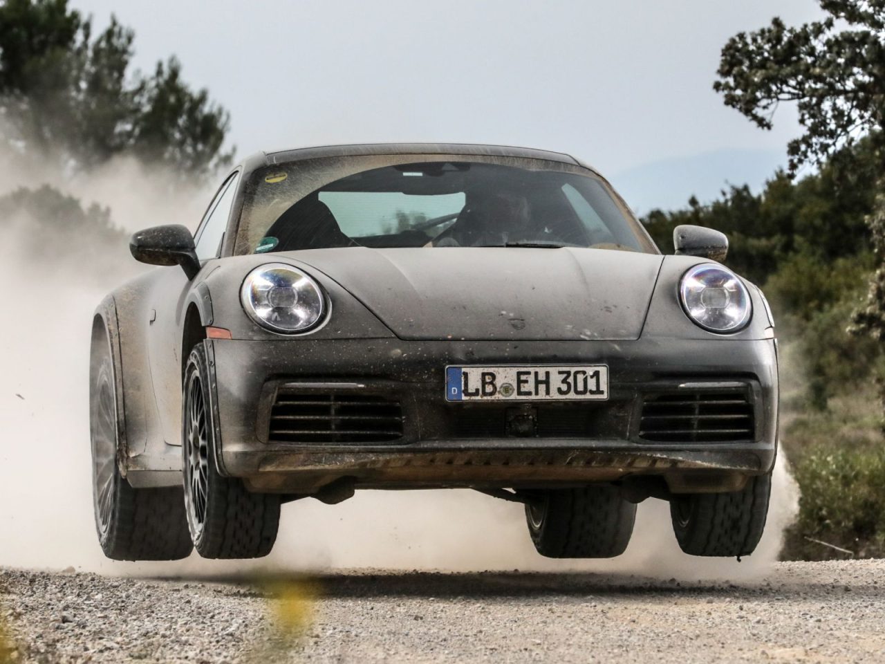 Een Porsche 911 Dakar met kenteken "LB EH 301" zweeft in de lucht en doet stof opwaaien tijdens het rijden op een onverharde weg in een landelijke omgeving.