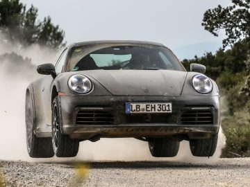 Een Porsche 911 Dakar met kenteken "LB EH 301" zweeft in de lucht en doet stof opwaaien tijdens het rijden op een onverharde weg in een landelijke omgeving.