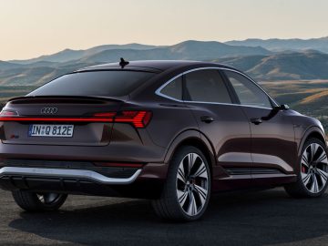 Een donkergekleurde Audi Q8 e-tron elektrische SUV staat geparkeerd op een verhard oppervlak met bergachtig terrein op de achtergrond onder een heldere hemel. Het achter- en zijprofiel van het voertuig zijn zichtbaar.