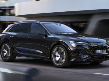 Een strakke zwarte Audi Q8 e-tron SUV rijdt op een stadsweg met moderne gebouwen op de achtergrond.