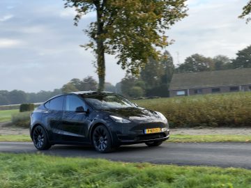 Een strakke Tesla Model Y Performance rijdt op een bewolkte dag over een landelijke weg met een boom en een huis op de achtergrond.