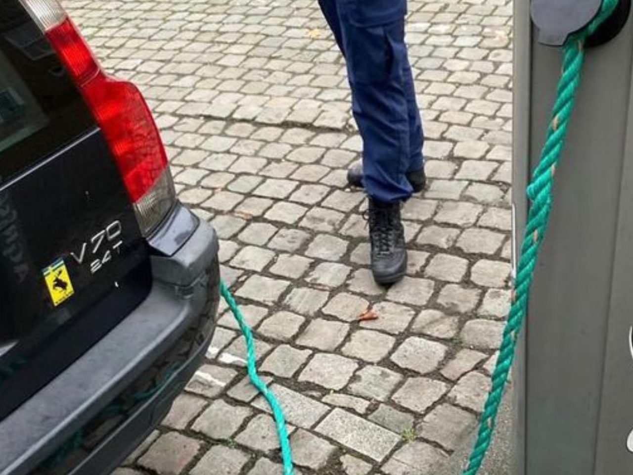 Op een geplaveide straat staat een persoon naast een Volvo die zijn elektrische batterij oplaadt, verbonden door een groene kabel.