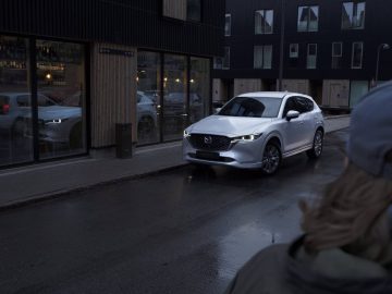 Een witte Mazda CX-5 SUV staat geparkeerd in een natte straat voor een gebouw met grote ramen. De koplampen van het voertuig zijn ingeschakeld en de reflectie ervan is zichtbaar in de ruiten. Een persoon met lang haar kijkt ernaar.