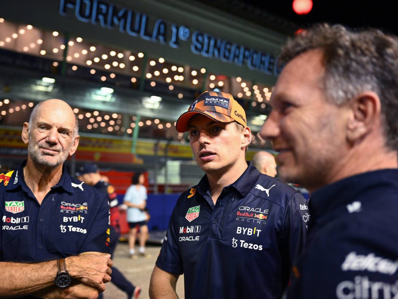 Drie mannen in teamkleding staan te praten voor een verlichte achtergrond van de Formule 1 in Singapore, waarbij de invloed van Adrian Newey duidelijk zichtbaar is in de technische discussies.