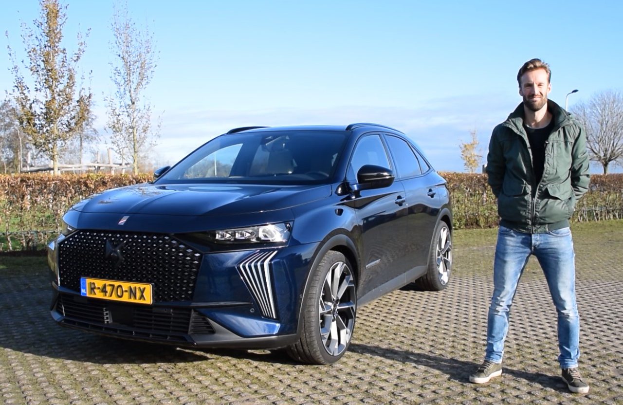 Een man staat op een zonnige dag buiten naast een geparkeerde donkerblauwe SUV met Nederlands kenteken en neemt misschien even de tijd om de opwinding van Vooruitblik Autojaar 2023 voor te bereiden.