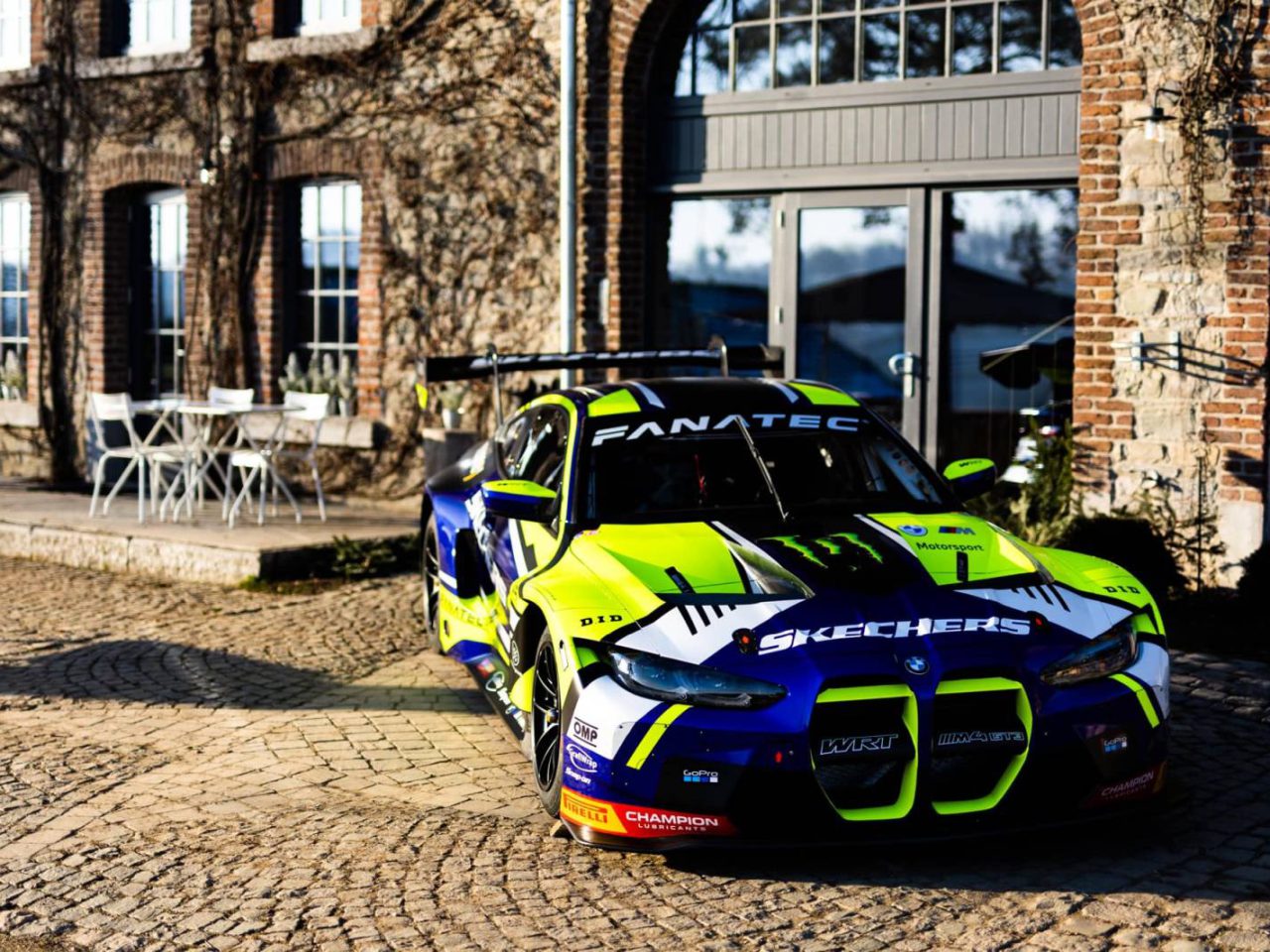 Een felgekleurde raceauto met sponsorlogo's, die doen denken aan de levendige stijl van Valentino Rossi, staat geparkeerd op een geplaveide ondergrond voor een bakstenen gebouw met boogramen.