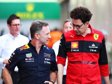 Twee mannen, één in een marineblauw shirt en één in een rood shirt, zijn met elkaar aan het praten terwijl ze buiten lopen. De scène weerspiegelt op subtiele wijze de kameraadschap die vaak te zien is in de Formule 1-paddocks, waar figuren als Christian Horner floreren. Op de achtergrond zijn nog drie andere personen te zien.