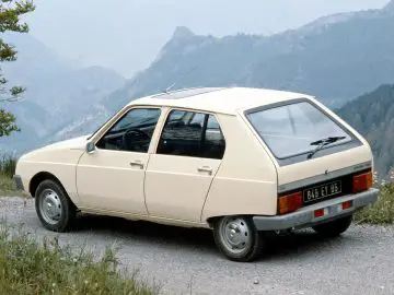 Een beige Citroën Visa hatchback staat geparkeerd langs een weg met bergachtig terrein op de achtergrond. De auto, bekend om zijn hoekige ontwerp, is vanuit driekwart achteraanzicht te zien.