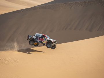 Off-road truck met sponsorlogo's vliegt in de lucht terwijl hij door zandduinen in een woestijn racet, wat doet denken aan een spannende Dakar-rally.