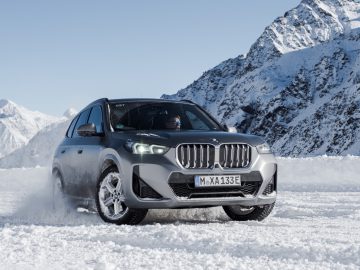 Een grijze BMW X1 SUV rijdt door besneeuwde bergen met een heldere hemel op de achtergrond.
