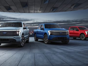 Drie elektrische pick-up trucks staan buiten geparkeerd in de schemering, een vooruitblik autojaar 2023. De trucks zijn wit, blauw en rood, zij aan zij opgesteld met de skyline van de stad zichtbaar op de achtergrond.
