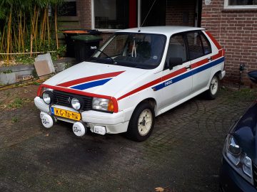 Op een stenen oprit staat een witte Citroën Visa hatchback met rode en blauwe racestrepen geparkeerd. Hij beschikt over vier ronde spots die op de voorbumper zijn gemonteerd.