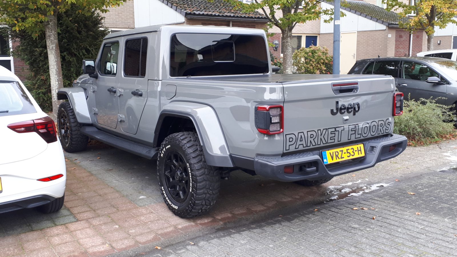 In een woonstraat staat een grijze Jeep Gladiator pick-up met een Nederlands kenteken en de advertentie "Parket Floors" op de achterklep geparkeerd.