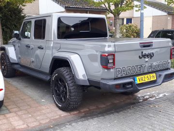 In een woonstraat staat een grijze Jeep Gladiator pick-up met een Nederlands kenteken en de advertentie "Parket Floors" op de achterklep geparkeerd.