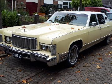 Een beige vintage Lincoln Continental sedan staat geparkeerd in een woonstraat met verspreide bladeren op het oppervlak en de grond. Op de achtergrond staan andere auto's geparkeerd.