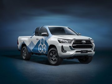 Een wit-blauwe Toyota Hilux pick-up met "H2" en "08" emblemen op de zijkant, geparkeerd tegen een donkerblauwe achtergrond.