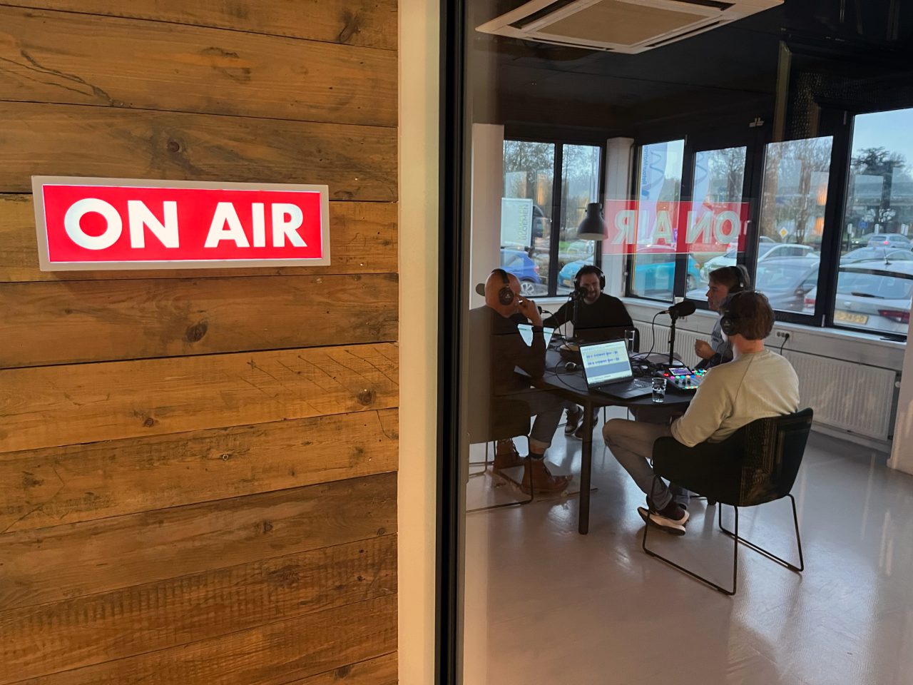 Een groep van vier mensen voert een discussie rond een tafel met microfoons en laptops in een opnamestudio. Aan de muur is een bordje met de tekst "ON AIR" zichtbaar, dat de opname van de eerste aflevering van de AutoRAI Podcast markeert.