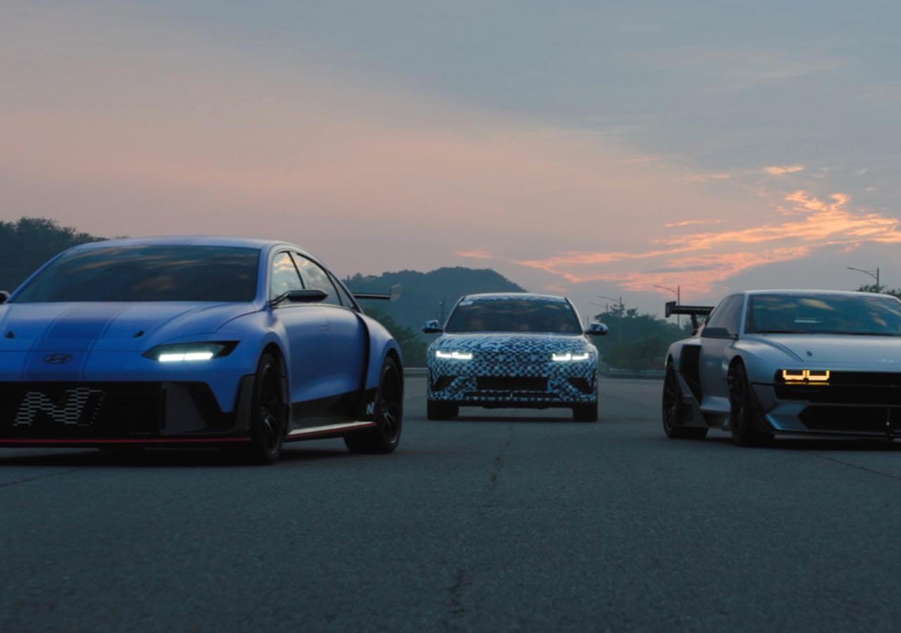 Drie sportieve auto's, een blauwe aan de linkerkant, een witte met blauwe patronen in het midden en een donkere Ioniq aan de rechterkant, rijden in de schemering over een open weg met een zonsondergang op de achtergrond.