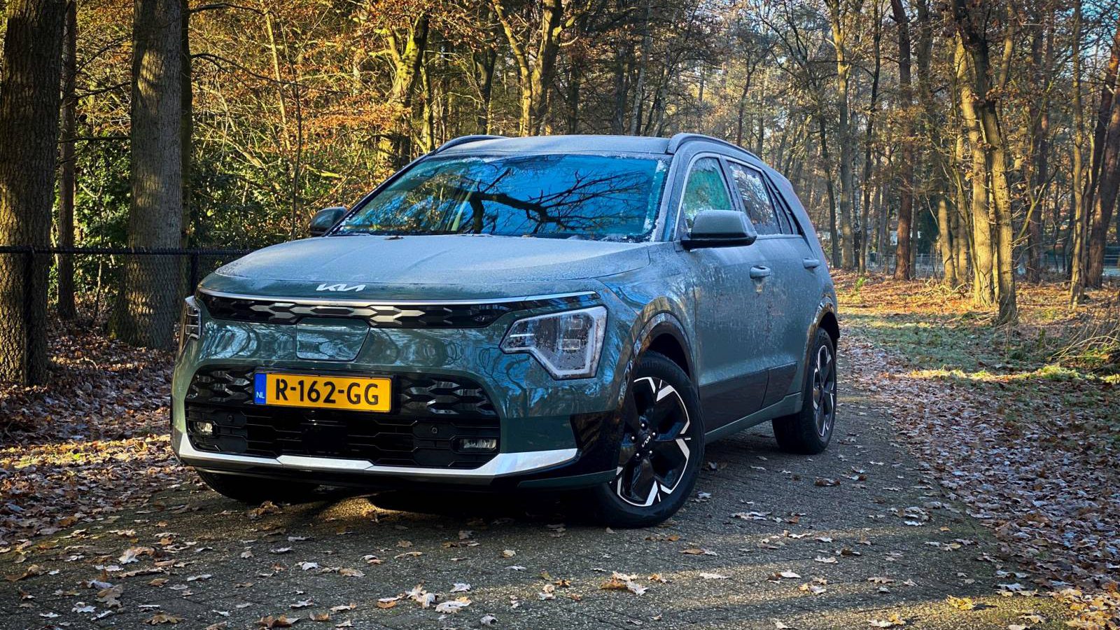 Een donkergroene Kia Niro EV staat geparkeerd op een bospad bedekt met gevallen bladeren. Het zonlicht filtert door de bomen en creëert een serene sfeer. Nederlands kenteken, R-162-GG, is zichtbaar.