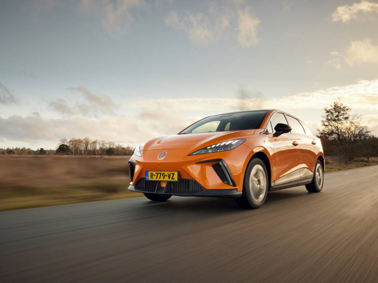 Oranje MG4 met Nederlands kenteken rijdend op een landweg, met een achtergrond van bomen en een gedeeltelijk bewolkte lucht.