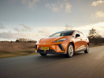 Oranje MG4 met Nederlands kenteken rijdend op een landweg, met een achtergrond van bomen en een gedeeltelijk bewolkte lucht.