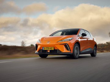 Een MG4 in een levendige oranje tint, met het kenteken "R-779-YZ", rijdt onder een bewolkte hemel over de weg.