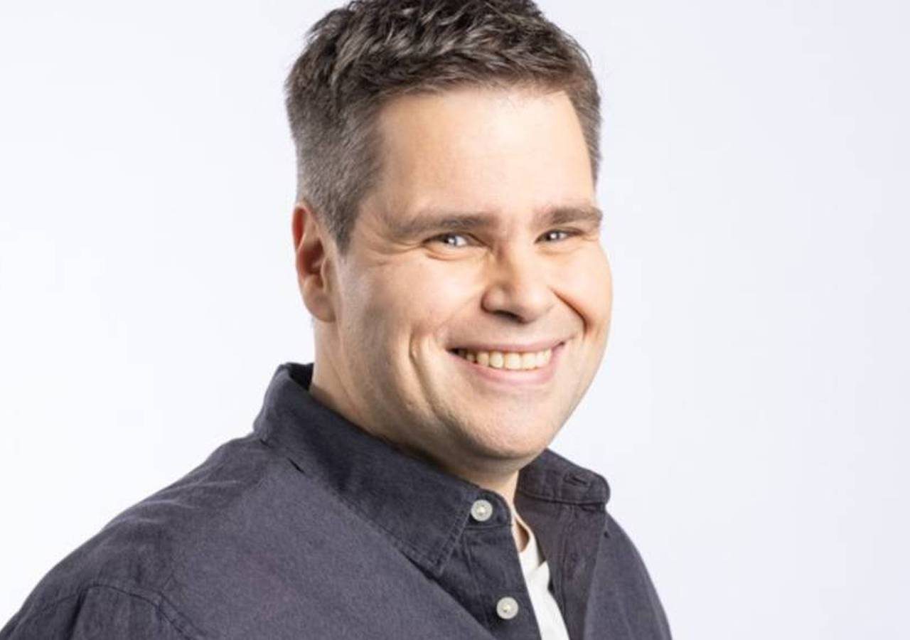 Nelson Valkenburg, met kort donker haar, lacht naar de camera, gekleed in een donker shirt over een wit t-shirt tegen een effen achtergrond.