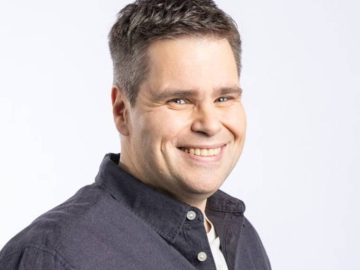 Nelson Valkenburg, met kort donker haar, lacht naar de camera, gekleed in een donker shirt over een wit t-shirt tegen een effen achtergrond.