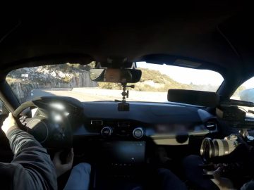 Binnenaanzicht van een auto met een rijdende persoon en een andere persoon die een camera op de passagiersstoel vasthoudt en de weg voor zich filmt. De weg buigt naar links met een bergachtig landschap op de achtergrond, dat doet denken aan een van Rosbergs spannende ritten.