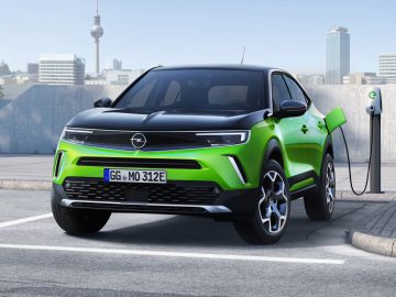 Een groen-zwarte Opel Mokka Electric staat geparkeerd en wordt opgeladen bij een station in een stedelijke omgeving met hoge gebouwen op de achtergrond.