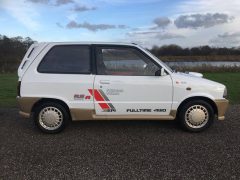 Zijaanzicht van een witte hot hatch met 'Fulltime 4WD'- en 'Turbo'-emblemen, geparkeerd op een geasfalteerde weg nabij een grasveld en een meer, onder een bewolkte hemel.