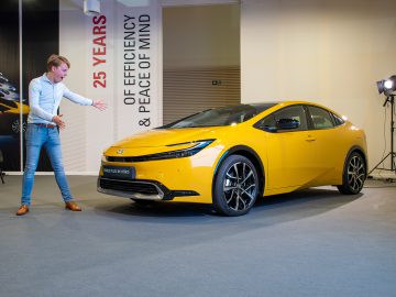 Een persoon gebaart naar een gele Toyota Prius 2023 Plug-in Hybrid die binnenshuis wordt tentoongesteld met een achtergrond ter ere van 25 jaar efficiëntie en gemoedsrust van het model.