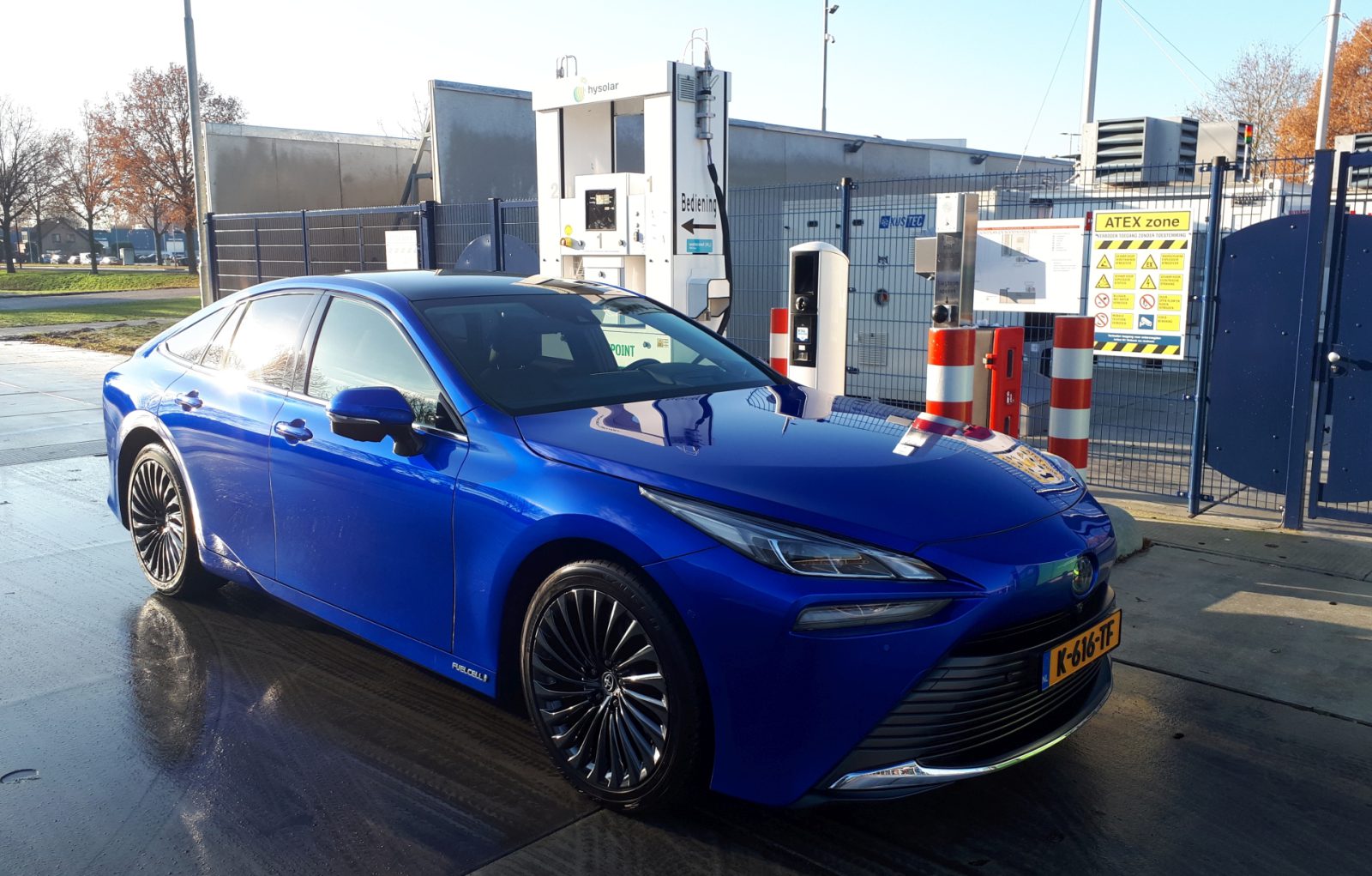 Bij een waterstoftankstation staat een blauwe sedan geparkeerd, met het mondstuk aangesloten op de brandstofinlaat van de auto, waarmee het proces van waterstoftanken wordt getoond.