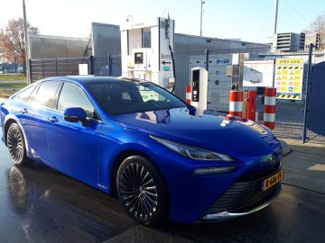 Bij een waterstoftankstation staat een blauwe sedan geparkeerd, met het mondstuk aangesloten op de brandstofinlaat van de auto, waarmee het proces van waterstoftanken wordt getoond.