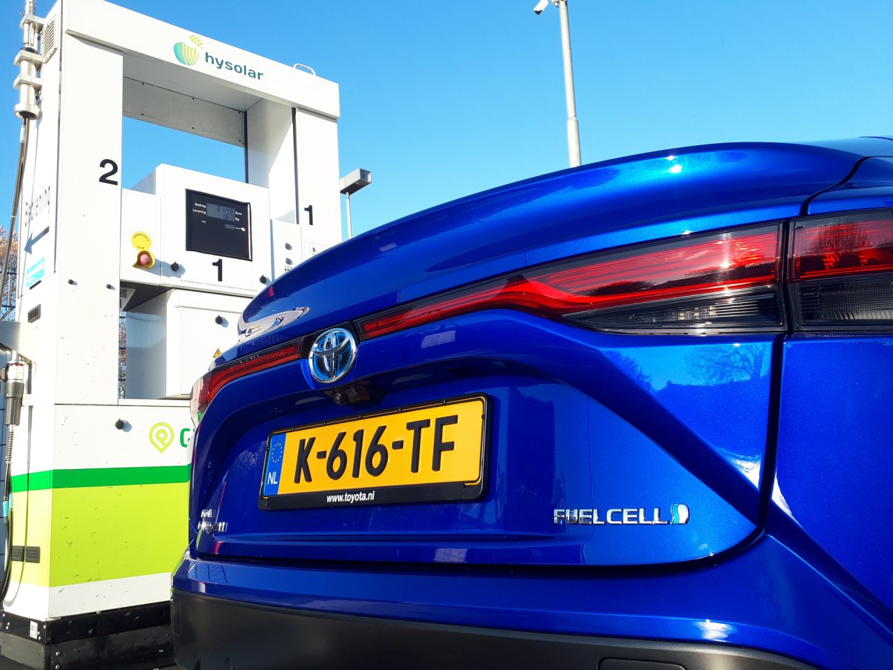 Een blauwe auto met geel kenteken K-616-TF, met het opschrift "FUELCELL", staat onder een helderblauwe hemel geparkeerd naast een Hysolar-tankstation, klaar voor waterstoftanken.