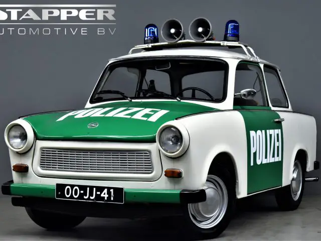 Een vintage Trabant P601 politieauto met "POLIZEI" geschreven op de deuren en motorkap. Het heeft een groen en wit kleurenschema, drie sirenes op het dak en een kentekenplaat met de tekst "00-JJ-41.