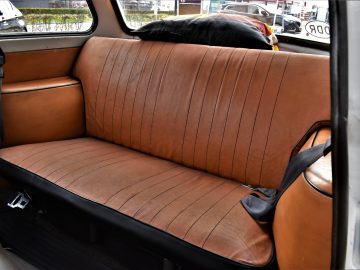Een bruinleren autostoeltje achterin met een zwarte veiligheidsgordel wordt getoond in wat lijkt op een Trabant P601. De stoel is voorzien van verticale stiksels en een hoofdsteun erbovenop. Door het autoraam kun je een glimp opvangen van geparkeerde auto's buiten.