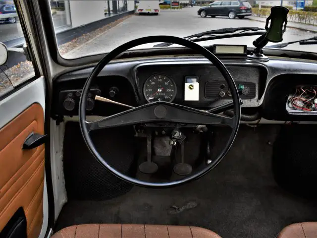 Binnenaanzicht van het dashboard van een oude Trabant P601 met een stuur, snelheidsmeter en pedalen; op het dashboard is een smartphone gemonteerd. Het zijpaneel aan de passagierszijde ontbreekt gedeeltelijk, waardoor de draden zichtbaar zijn.