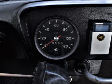 Een close-up van een Trabant P601-dashboard met een zwarte snelheidsmeter die een stand van 89,06 km/u weergeeft en een vintage sigarettenaanstekerhouder aan de rechterkant.