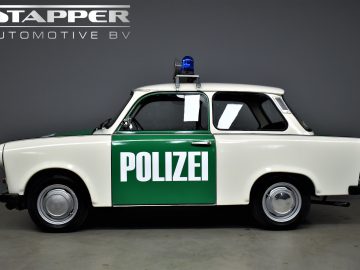 Zijaanzicht van een vintage witte en groene Trabant P601 politieauto met het woord "POLIZEI" op de deur en een blauwe sirene erop. In de linkerbovenhoek is het logo "STAPPER AUTOMOTIVE BV" zichtbaar.