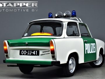 Een witte en groene vintage Trabant P601 politieauto met de opdruk "POLIZEI" wordt tentoongesteld in een showroom. Op het kenteken staat "00-JJ-41." De afbeelding is voorzien van het logo "STAPPER AUTOMOTIVE BV".