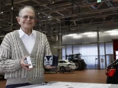 Een oudere man in een trui staat in een industriële ruimte, glimlachend naar de camera terwijl hij een doos vol cilindervormige voorwerpen vasthoudt. De scène legt de essentie vast van een uitvinder aan het werk. Op de achtergrond zijn verschillende voertuigen zichtbaar, wat bijdraagt aan de inventieve sfeer.