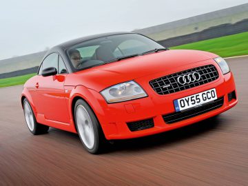 Een rode Audi TT Coupé snelt over het circuit met een onscherpe achtergrond en op het kenteken staat "OY55 GCO". De autowaarde van dit stijlvolle voertuig blijkt duidelijk uit de gestroomlijnde prestaties en het opvallende ontwerp.