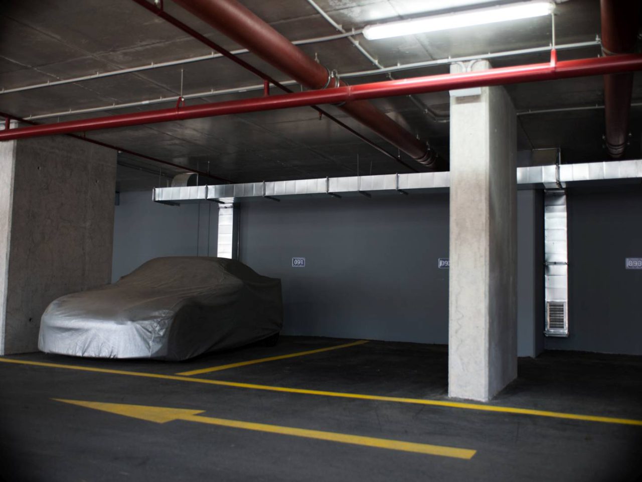 Een overdekte auto staat geparkeerd in een autostalling in een overdekte parkeergarage met grijze muren, betonnen pilaren en bovenleidingen. Op de vloer zijn gele pijlmarkeringen zichtbaar.